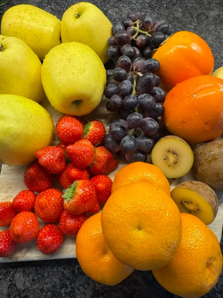 Eine bunte Auswahl an frischen Früchten auf einem Holzbrett, darunter gelbe Äpfel, dunkle Weintrauben, Kaki, Erdbeeren, Orangen und halbierte Kiwi, alle mit Wassertropfen bedeckt.