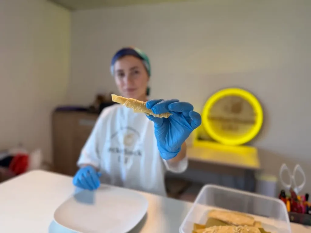 Eine Mitarbeiterin von LeckerSnack LILI mit blauen Handschuhen und Haarnetz hält ein Stück Pastila in die Kamera. Im unscharfen Hintergrund ist ein gelb leuchtendes Logo sichtbar.