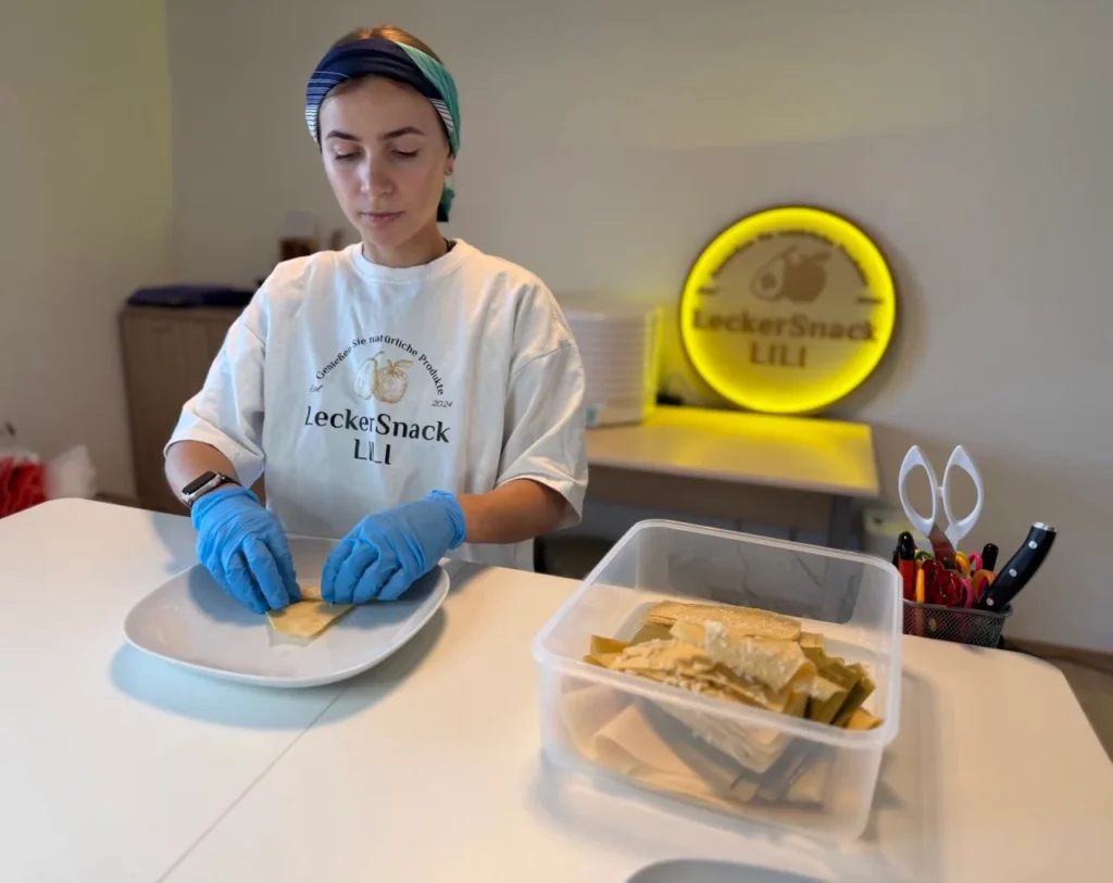Eine Mitarbeiterin von LeckerSnack LILI in Schutzkleidung (Haarnetz, blaue Handschuhe) hält ein aufgerolltes Stück dunkle Pastila in die Kamera, im Hintergrund das leuchtende gelbe Markenlogo.